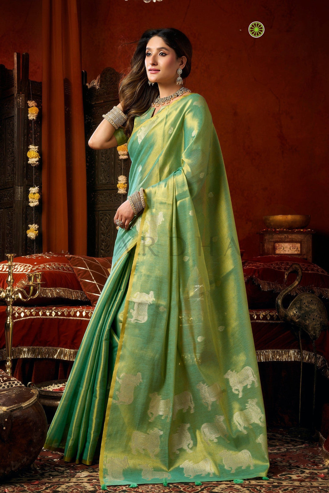 Woman in a green saree with a traditional setting Side standing view of the sea green festive wear heavy tissue saree highlighting the traditional Pichwai-style cow motifs and flawless drape.