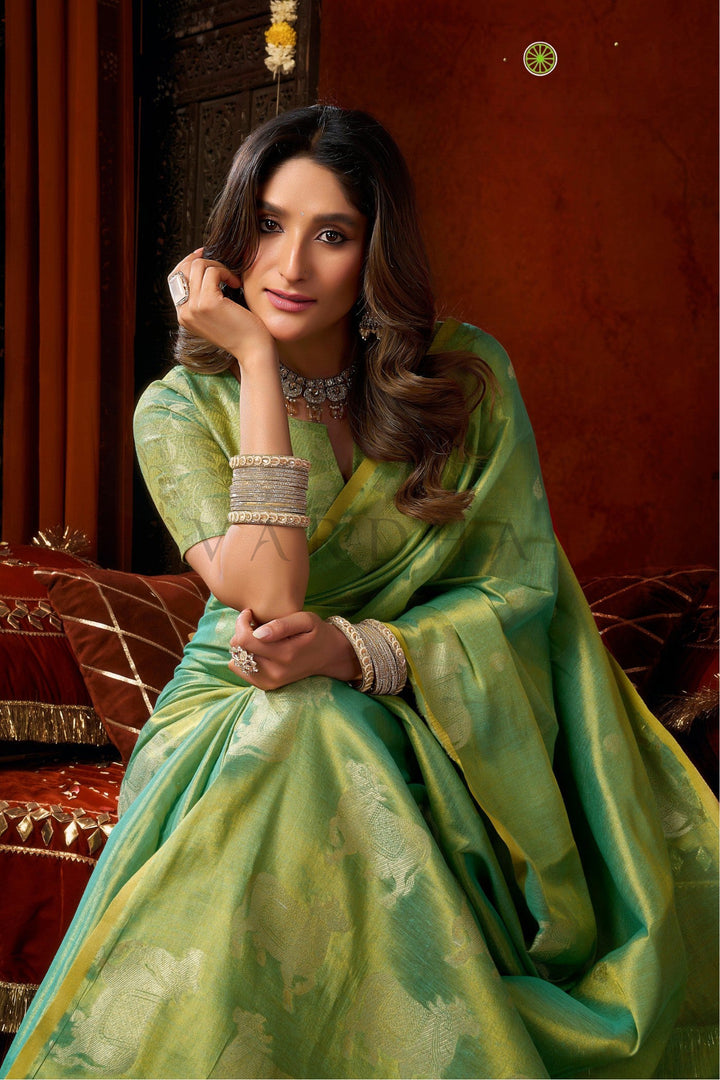A woman wearing a green silk saree with subtle floral motifs, seated in an ornate traditional setting with warm brown walls and red cushions. Beautiful woman sitting gracefully in a vibrant mint green tissue silk saree, showcasing the luxurious drape and matching woven blouse.
