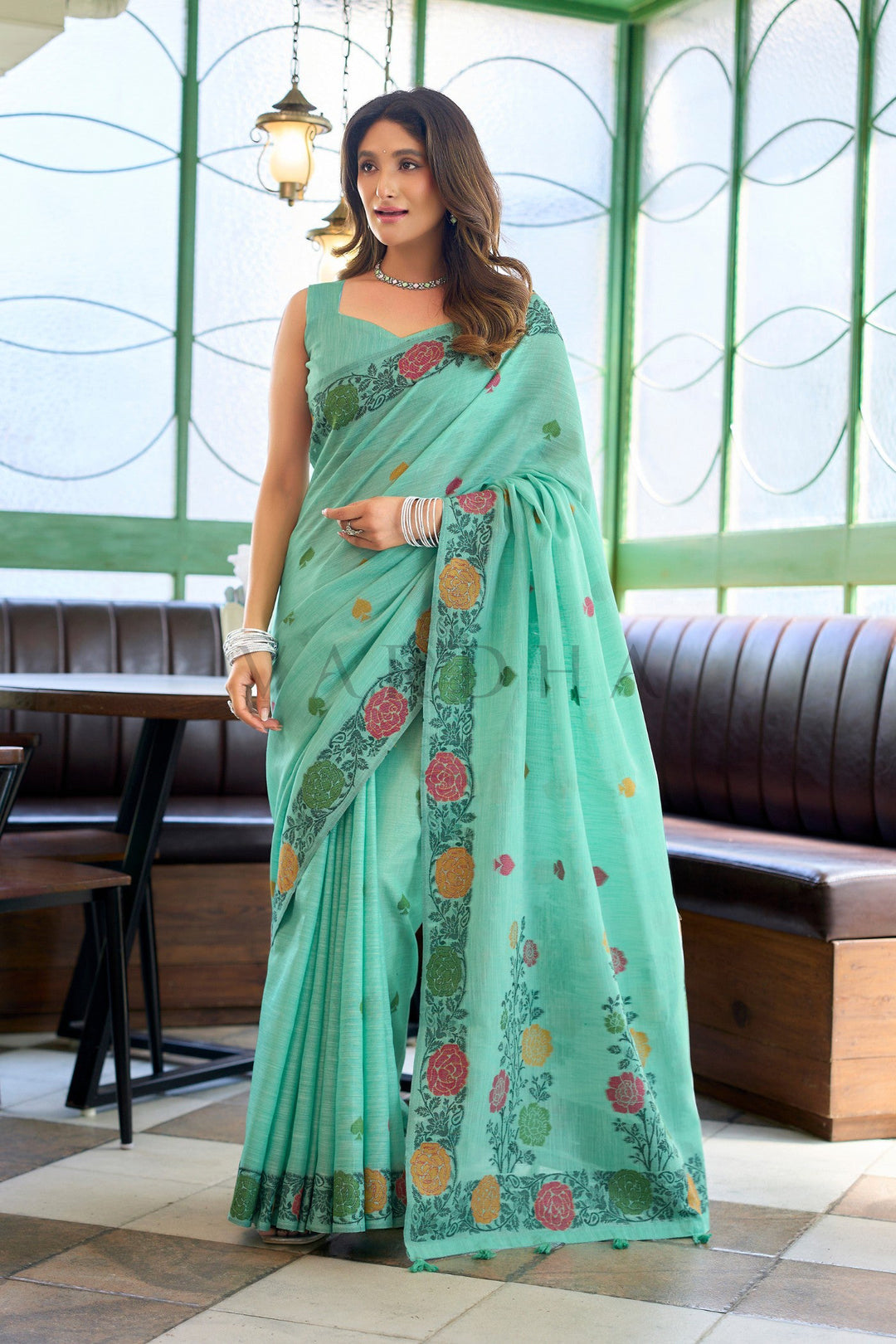 Woman wearing a light blue saree with floral patterns in an indoor setting. Full-length view of a woman wearing an elegant mint green linen jacquard handloom saree with a multi-colored woven floral border and fancy tassels.