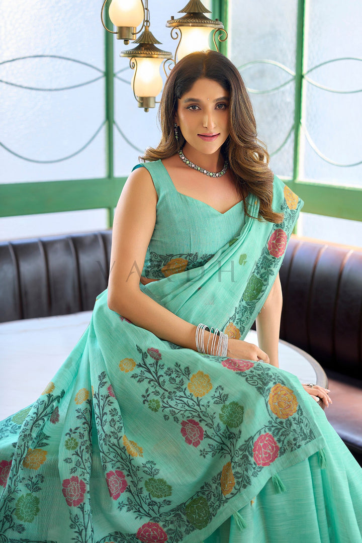 A woman wearing a mint green saree with colorful floral prints, seated in an indoor setting with a dark leather sofa and hanging lights in the background. Beautiful woman sitting elegantly in a sea green linen jacquard saree, showcasing the comfortable drape and exquisite floral woven pallu design.