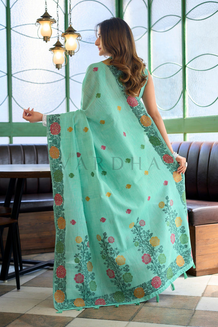 Woman holding a light blue saree with floral patterns in a room with green walls and a window. Back view of the green linen jacquard saree highlighting the stunning oversized woven floral vines and matching fancy tassels on the pallu.