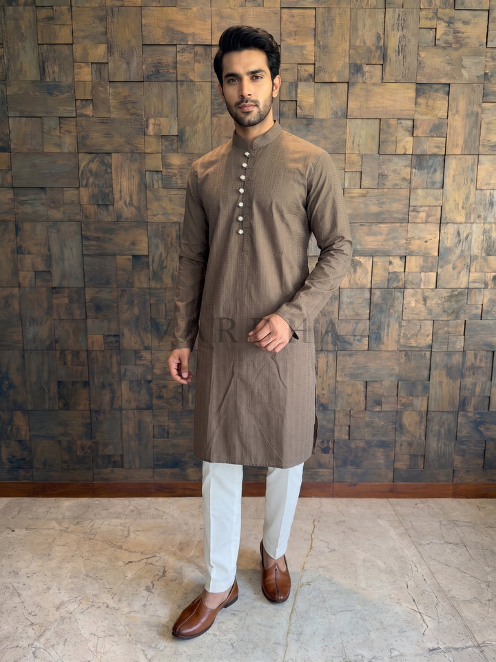 Man wearing a brown kurta with white pants against a wooden paneled wall. Men’s Mocha Brown cotton kurta featuring a woven vertical micro-stripe (dobby) texture that adds visual depth and elongates the silhouette.