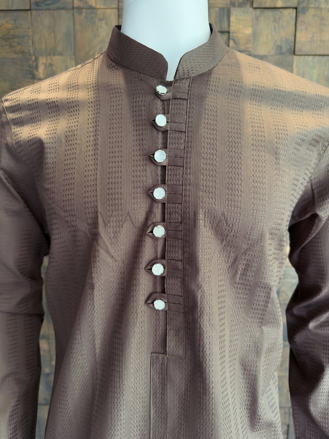 Brown kurta with white buttons on a mannequin against a stone wall background. Close-up of a designer coffee-brown kurta highlighting the extended chest placket with traditional fabric loops and striking metallic silver mirror-finish buttons.