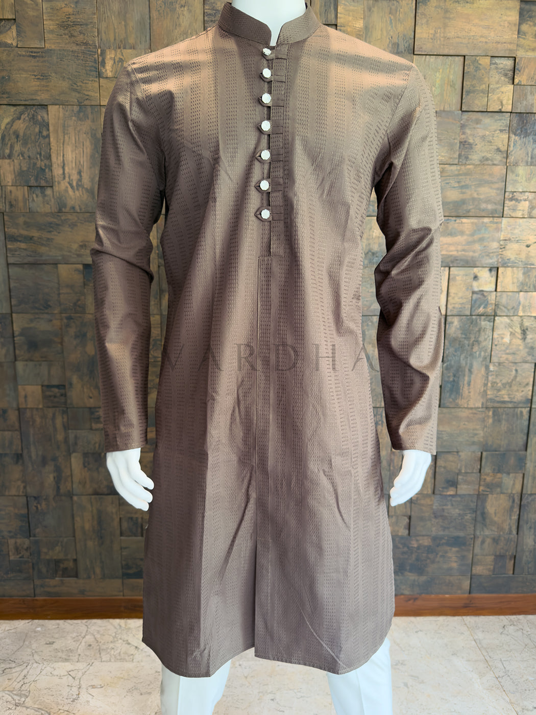 Brown kurta on a mannequin against a wooden paneled wall. A knee-length Mocha Brown kurta featuring an extended placket with fabric tabs and high-shine silver buttons for a bold, modern contrast.