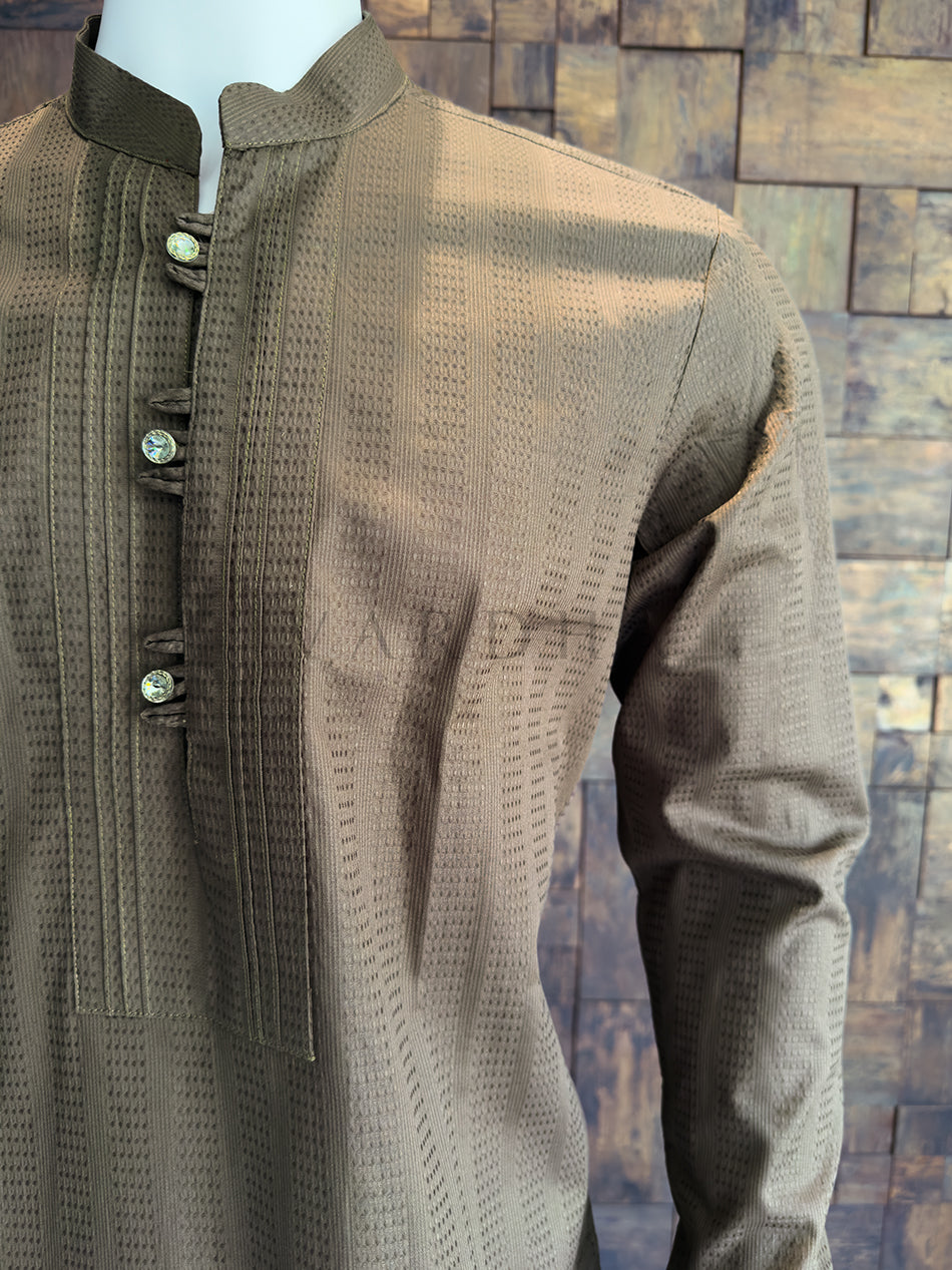 Brown textured shirt on a mannequin against a wooden paneled wall. tailored with a clean Mandarin collar. The centerpiece of the design is the meticulously crafted chest placket. It is framed by sharp, vertical pleats (pintucks) that add a formal, structured "bib" effect. The placket itself is secured with traditional fabric button loops (tabs) and finished with premium, crystal-studded designer buttons that provide a luxurious touch of shine against the matte brown fabric.