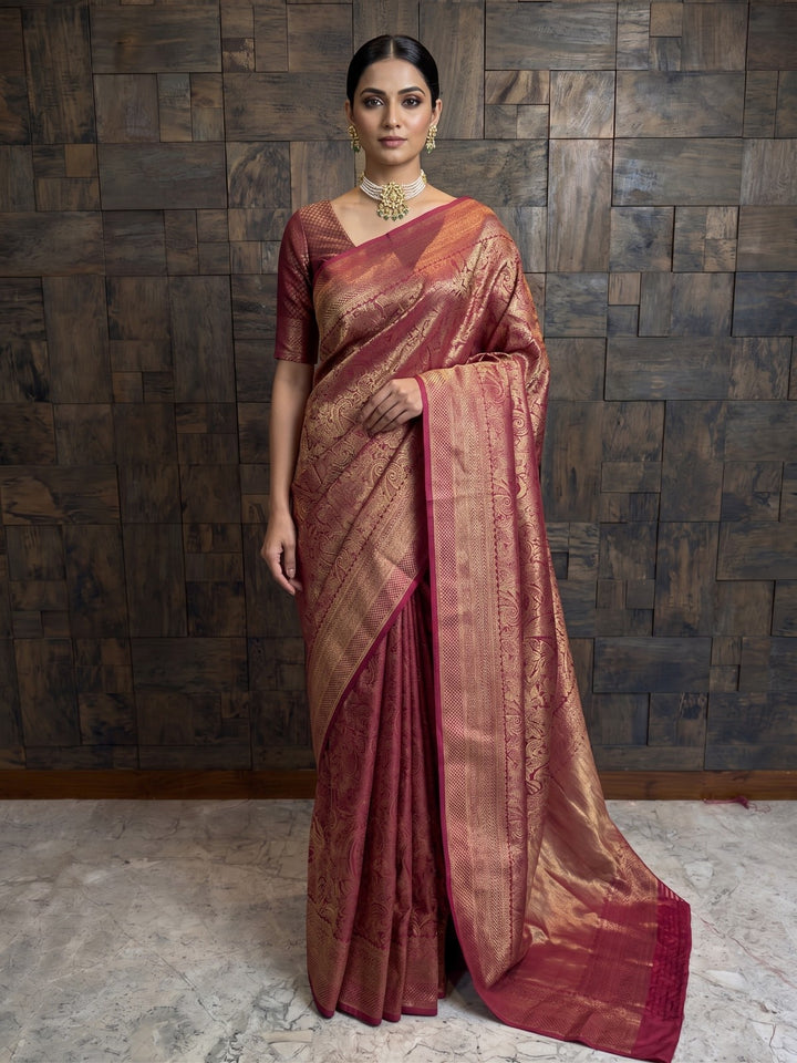 A full-length front view of a woman standing and posing in a rich mulberry purple Kanjivaram silk saree, heavily embellished with intricate gold zari floral and traditional motifs. She is wearing a matching short-sleeved blouse, a pearl choker necklace with matching earrings, and her hair is styled in a neat low bun.