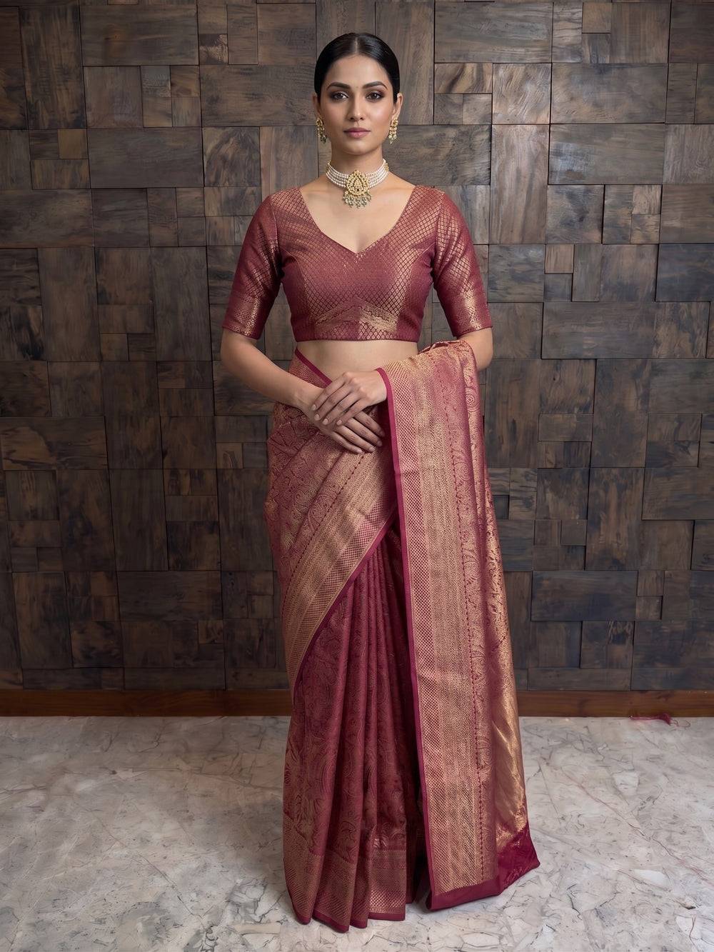 A full-length front view of a woman wearing a mulberry purple Kanjivaram silk saree. She has one hand resting gently against her waist, showcasing the elegant drape, the wide gold zari borders, and the dense weaving patterns across the fabric.