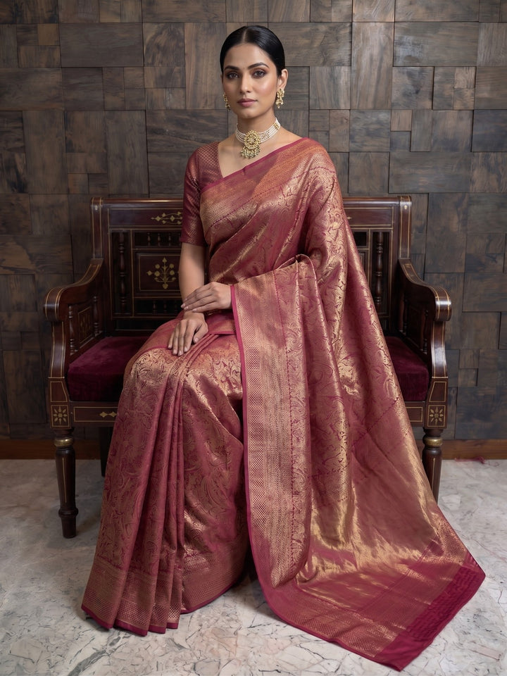 A full-length shot of a woman seated gracefully on an ornately carved wooden bench with maroon upholstery. She is wearing a mulberry purple Kanjivaram silk saree, which is beautifully draped around her, highlighting the radiant gold zari work against the dark wood background.
