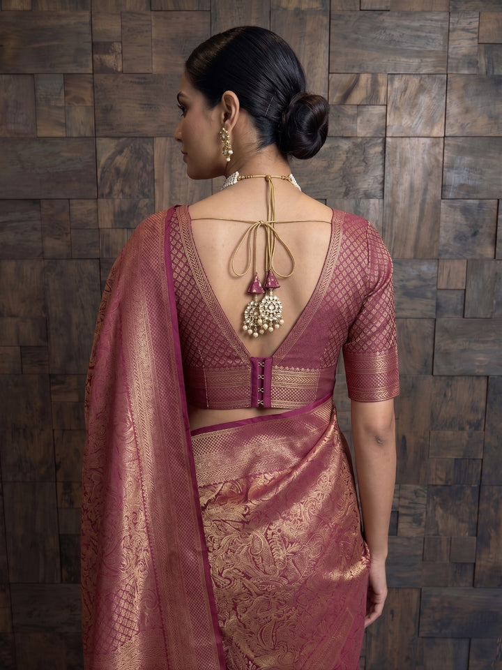 A detailed back view focusing on the upper half of a woman in a mulberry purple Kanjivaram silk saree. The matching blouse features a deep V-neck back, a row of closures at the lower waist, and a top string tie adorned with large, ornate, jewelry-like tassels.