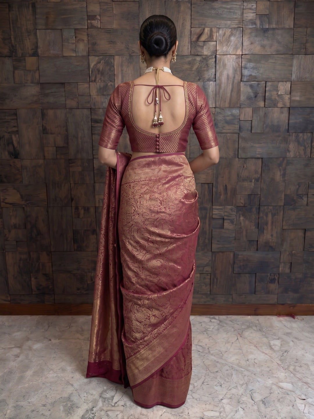 A full-length back view of a woman wearing a mulberry purple Kanjivaram silk saree, holding the edge of the pallu slightly to the side. Her blouse showcases a distinct circular keyhole cutout on the back, complete with a top string tie, tassels, and decorative buttons at the base