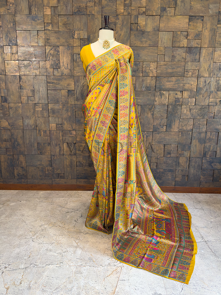 Yellow saree with colorful patterns on a mannequin against a wooden paneled wall. Full-length view of a vibrant mustard yellow silk saree draped on a mannequin, featuring exquisite multi-colored Meenakari weaving, all-over geometric patterns, and intricate traditional borders. Ideal for Indian weddings and festive ethnic occasions.