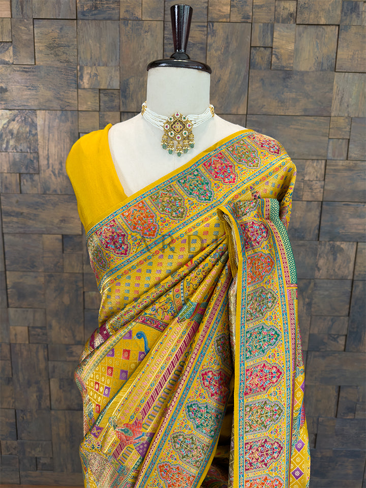 Colorful saree with intricate patterns on a mannequin against a wooden paneled wall. Close-up of a traditional yellow bridal silk saree showing a draped pallu over the shoulder, adorned with woven peacock, floral, and diamond lattice Meenakari designs in vibrant blue, pink, and green threads.