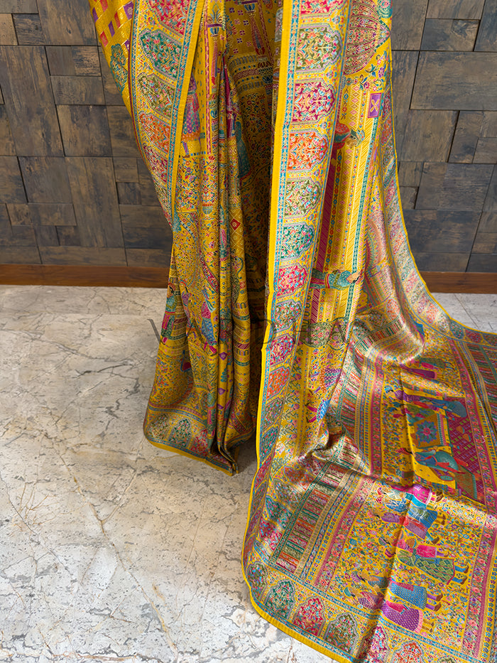 Colorful patterned fabric draped over a wooden surface Lower drape of a heavy heritage woven silk saree highlighting the rich, multi-colored border and traditional motifs, including woven human figures and intricate floral vines cascading elegantly toward the floor.