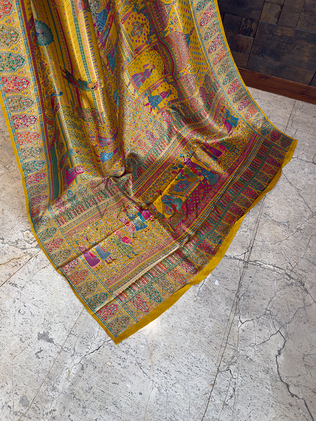 Colorful patterned fabric on a textured floor Trailing edge of a luxurious yellow festive saree resting on a stone floor, displaying an elaborate pallu woven with intricate royal court figures, peacock motifs, and ornate traditional Indian patterns.
