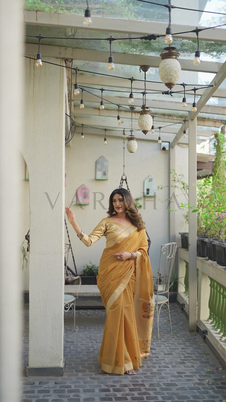A woman wearing a mustard yellow saree with a matching blouse stands on a covered outdoor walkway. The setting features a white wall, hanging string lights, decorative lanterns, and potted plants. Beautiful model posing gracefully on a decorated balcony, showcasing the perfect drape and vibrant golden hue of the mustard cotton jacquard festive saree.