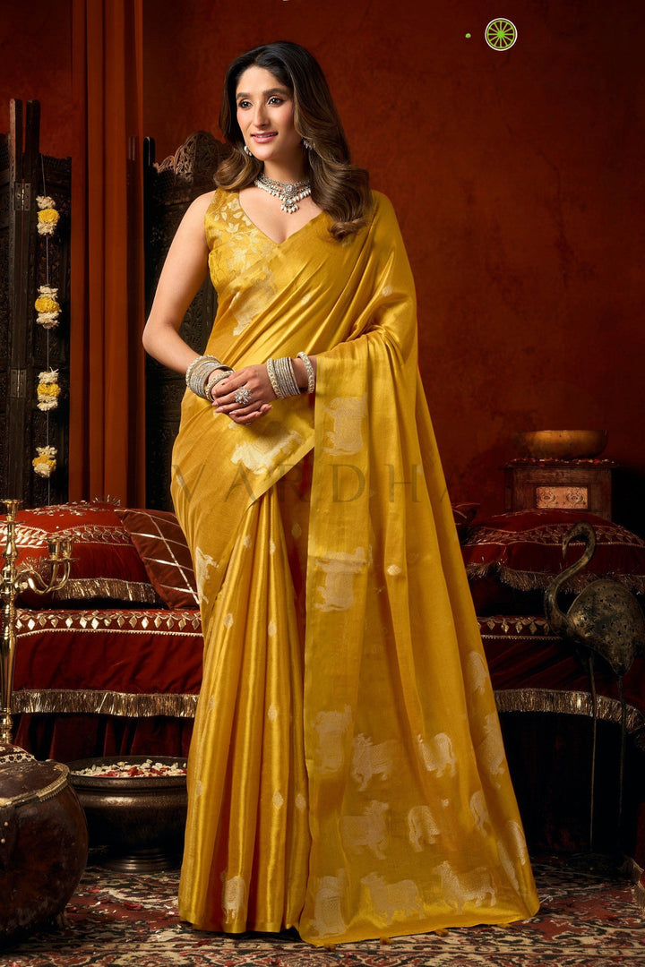 A woman wearing a golden yellow silk saree with white zari motifs, standing in a traditional Indian room with red and gold decor. Full front view of a traditional mustard yellow heavy tissue saree featuring allover silver woven cow buttas and a rich golden zari border.