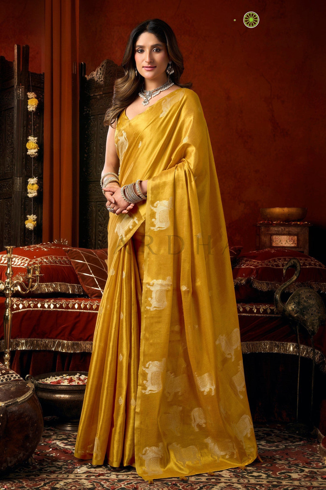 Woman in a yellow saree with a traditional setting Side standing view of the mustard yellow festive wear heavy tissue saree highlighting the traditional Pichwai-style cow motifs and flawless drape.