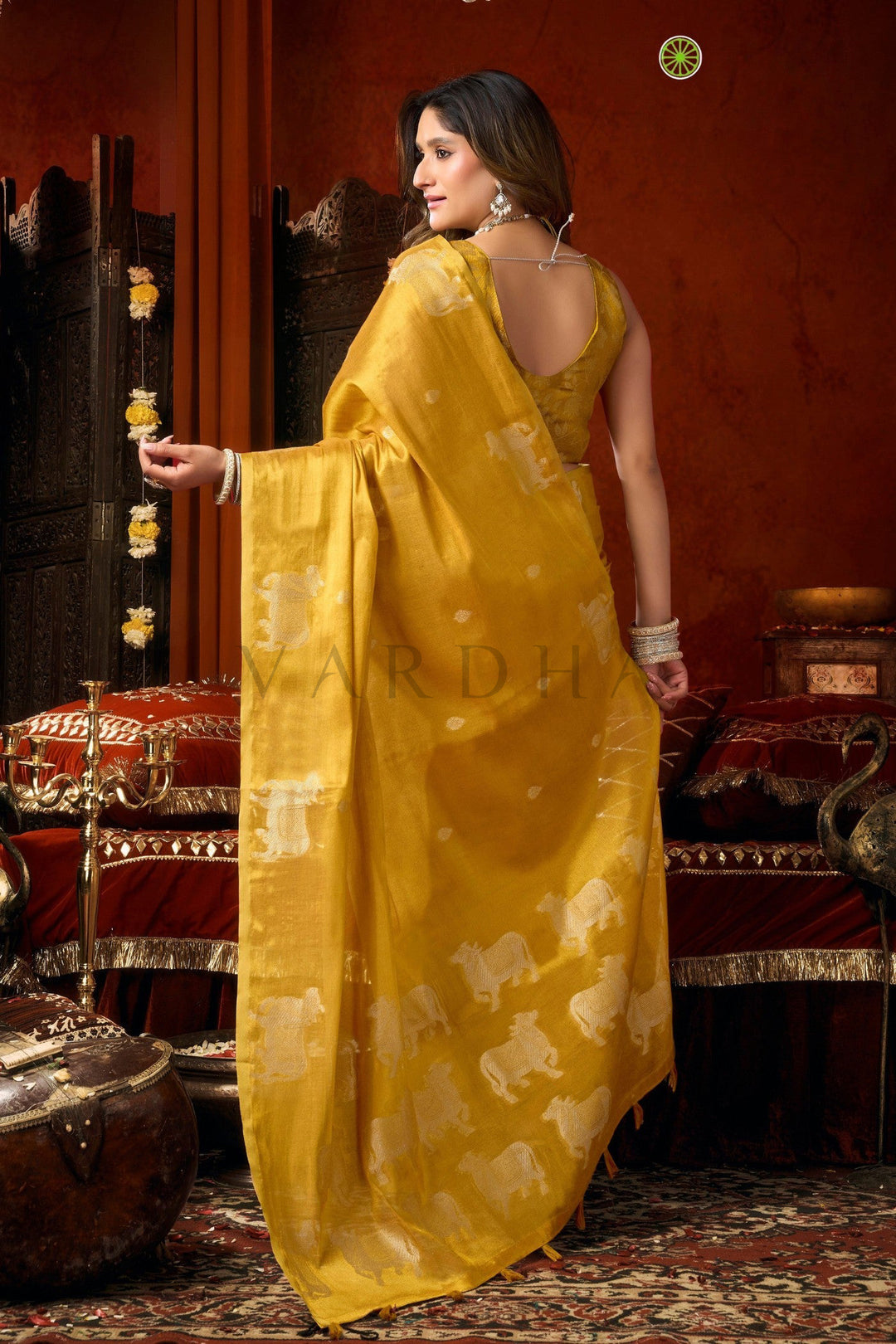 A woman wearing a mustard yellow saree with white elephant motifs, standing in a traditional Indian room with red walls and ornate furniture. Back view of the radiant golden yellow tissue saree, emphasizing the rich metallic sheen, woven silver cow designs, and elegant fancy tassels.