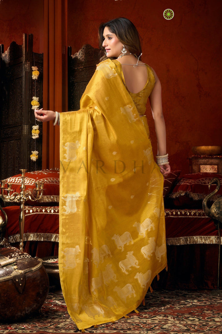 A woman wearing a mustard yellow saree with white elephant motifs, standing in a traditional Indian room with red walls and ornate furniture. Back view of the radiant golden yellow tissue saree, emphasizing the rich metallic sheen, woven silver cow designs, and elegant fancy tassels.