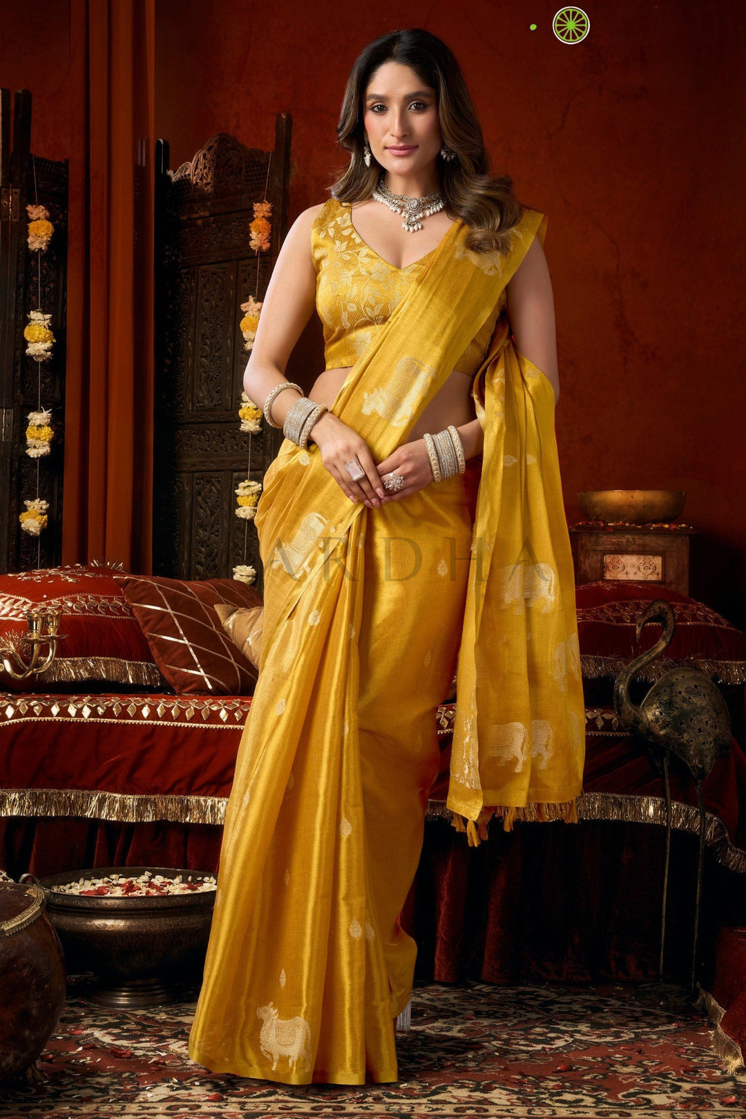 A woman wearing a bright yellow silk saree with subtle white motifs, standing in a richly decorated traditional room with red walls and ornate furniture. Stylish front drape of the mustard yellow heavy tissue silk saree showcasing its luxurious texture, perfect for wedding wear and festive occasions.