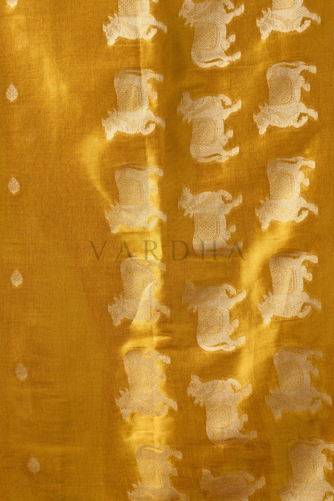Textured fabric with a pattern of small shapes on a yellow background Detailed fabric close-up of the golden yellow heavy tissue saree showcasing the intricate silver zari woven cow (Pichwai) buttas and fancy tassels.