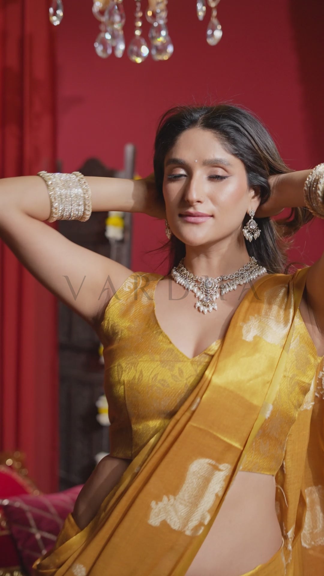A woman wearing a golden yellow saree with white floral patterns, styled with a matching blouse, silver jewelry, and bangles, posing in front of a red curtain background. Glamorous evening shot of a woman tying her hair while wearing the vibrant mustard yellow heavy tissue saree, highlighting its regal metallic glow.