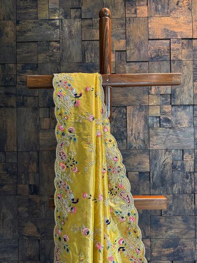 Close-up of mustard tissue saree featuring multi-color floral and bird embroidery with sequins and zari highlights.