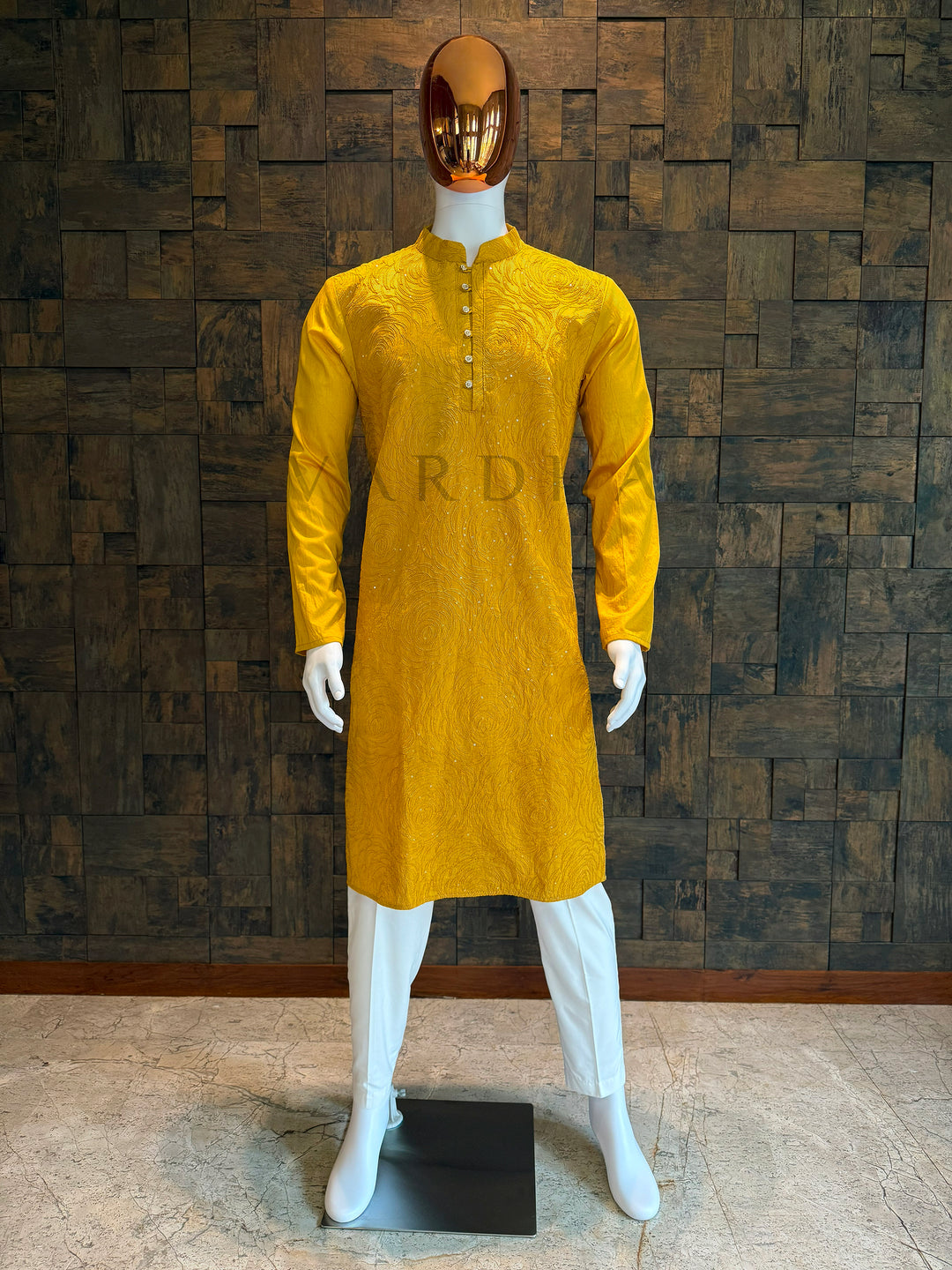Yellow kurta on a white mannequin against a textured brown wall. Marigold yellow viscose men’s kurta with mandarin collar and rosette swirl embroidery accented by sequins, displayed on a mannequin with white churidar—perfect for festive and wedding events.