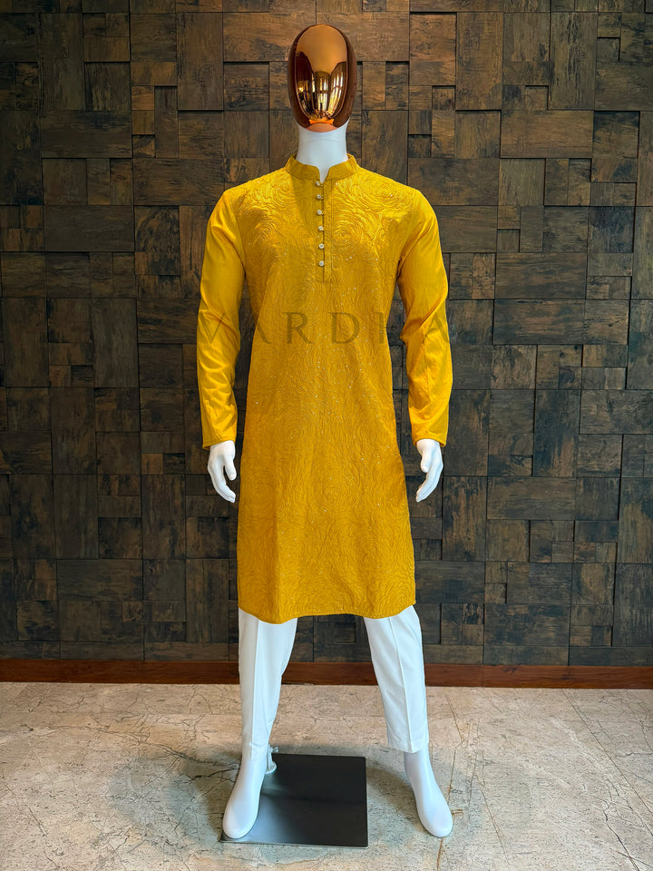 Yellow kurta on a white mannequin against a textured brown wall. Marigold yellow viscose men’s kurta with mandarin collar and rosette swirl embroidery accented by sequins, displayed on a mannequin with white churidar—perfect for festive and wedding events.