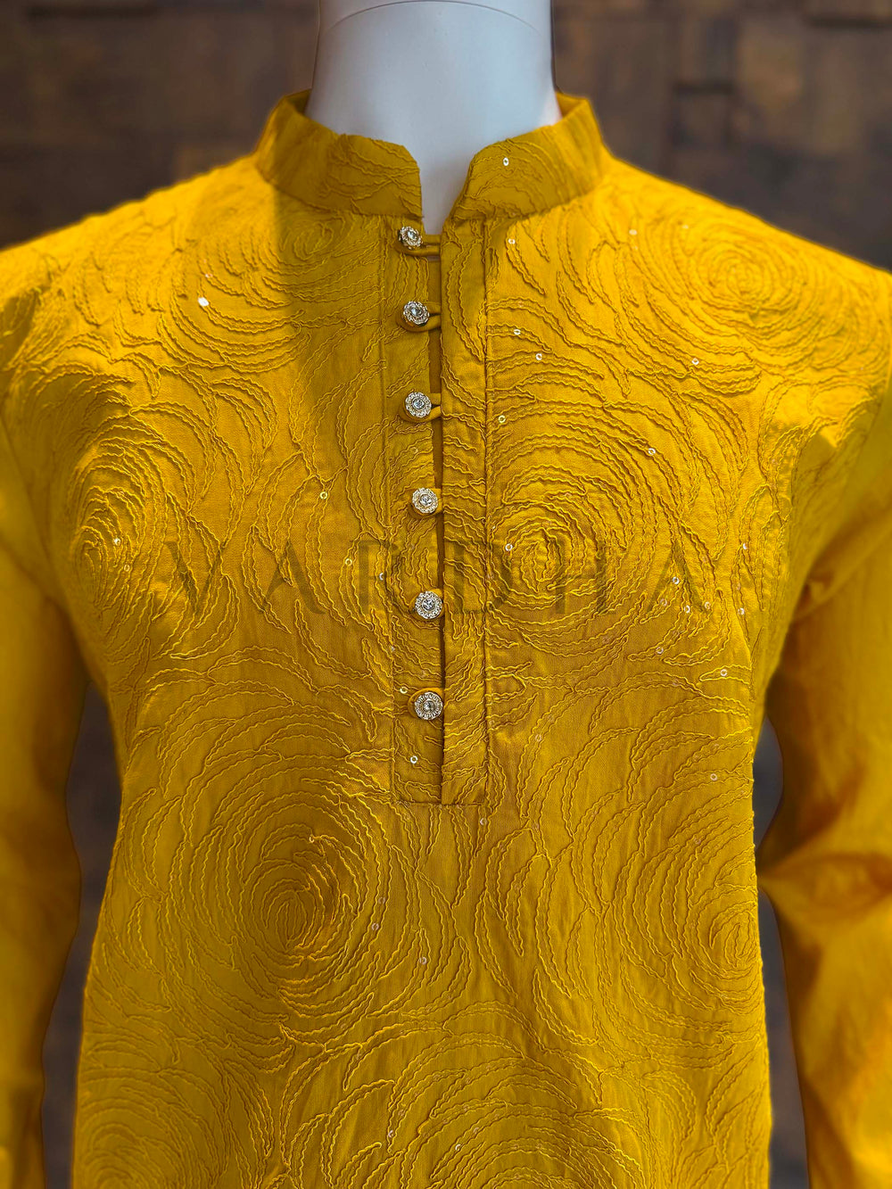 Yellow embroidered shirt on a mannequin against a dark background Close-up of marigold kurta neckline showing band collar and ornate buttons with tonal rosette embroidery and subtle sequin sparkle on smooth viscose fabric.