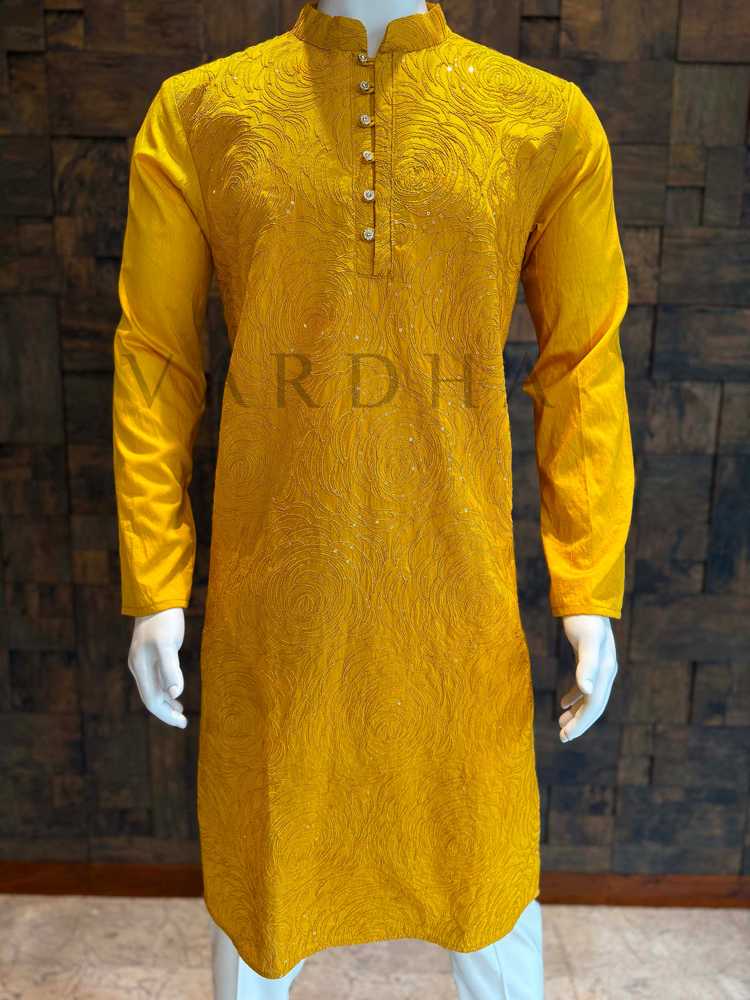 Yellow kurta on a mannequin against a wooden paneled wall. Full-length view of yellow embroidered kurta with long sleeves and curved hem, styled for haldi, mehendi and sangeet celebrations. Macro texture of viscose kurta capturing layered rosette stitches, fine sequin dots and gentle fabric sheen that photographs beautifully.