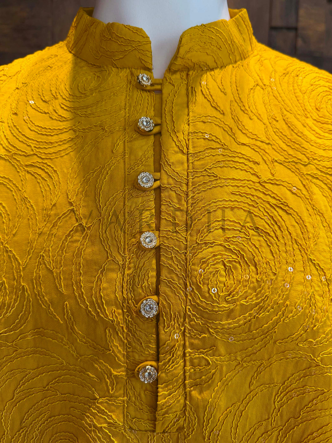 Yellow embroidered jacket with buttons on a mannequin against a dark background Front chest detail highlighting concentric swirl threadwork and scattered micro-sequins on marigold viscose, framing a neat button placket for elegant menswear.