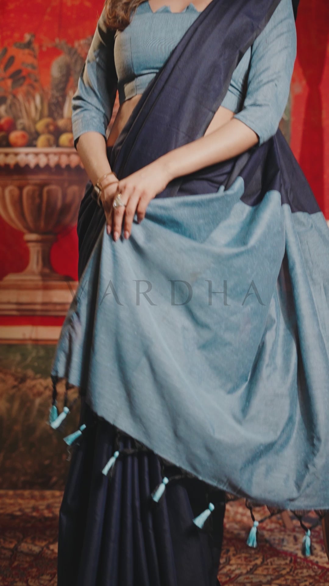 A woman wearing a light blue and navy blue saree with matching blouse, standing in front of a red decorative background. The image has a faint watermark text &