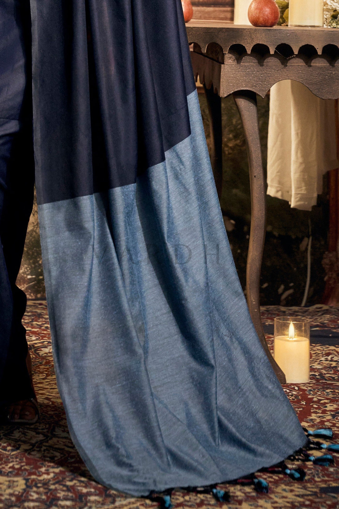 Blue fabric draped over a chair with a candle and decorative items in the background Detailed fabric close-up of the navy blue mal cotton saree highlighting the perfectly pleated texture and the powder blue contrast pallu.