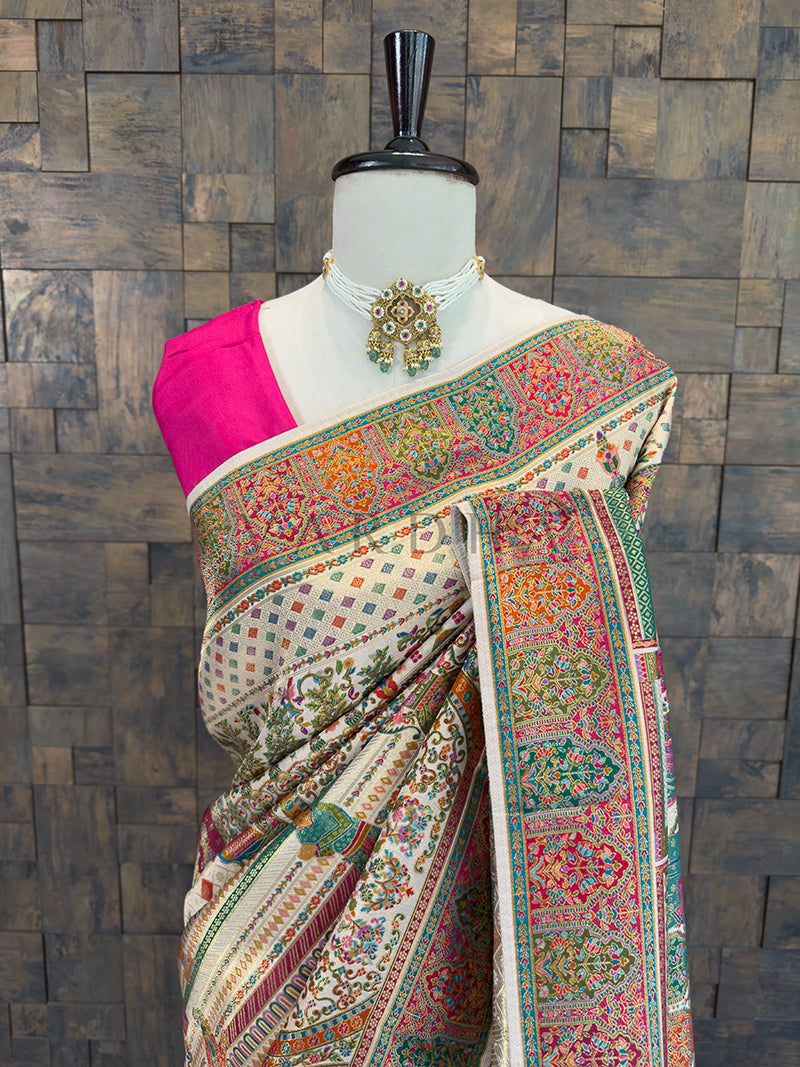 Colorful saree with intricate patterns on a mannequin against a wooden paneled wall. Close-up of an ivory traditional bridal silk saree with a vibrant pink blouse, highlighting the shoulder drape adorned with multi-color Meenakari floral vines and geometric diamond weaves, accessorized with a classic pearl choker.