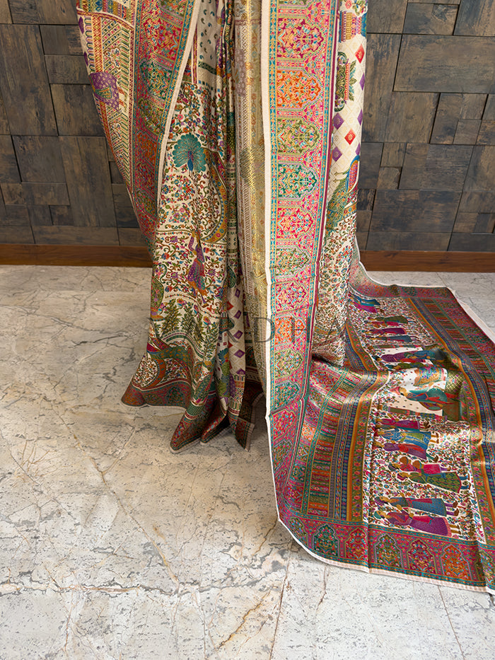 Colorful patterned fabric draped over a chair against a wooden paneled wall. Lower drape of a heavy off-white heritage woven silk saree, showcasing the exquisite multi-colored borders with intricate peacock, floral, and traditional human figure motifs cascading elegantly toward the floor.