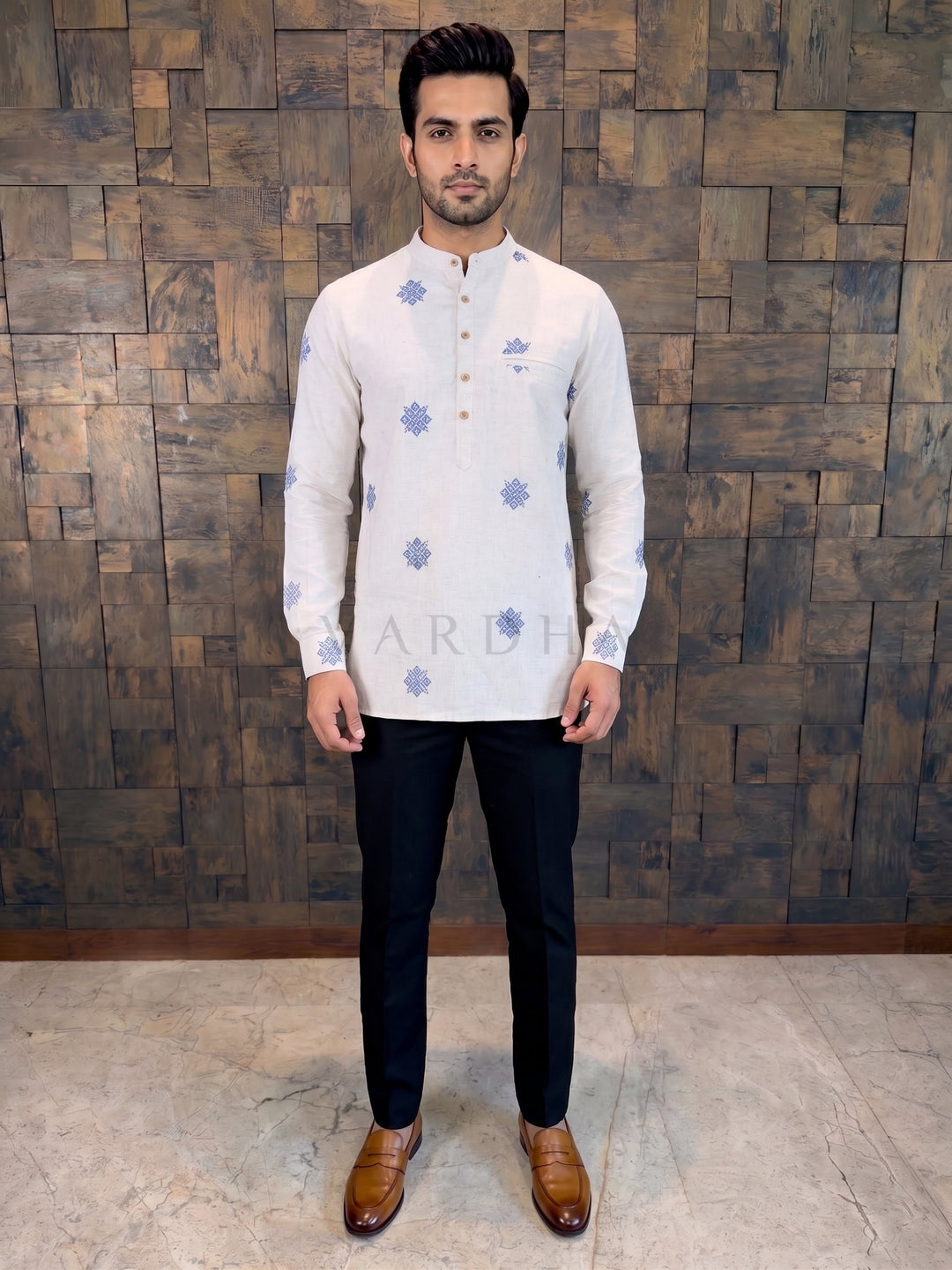 Man wearing a white kurta with blue patterns against a wooden paneled wall.