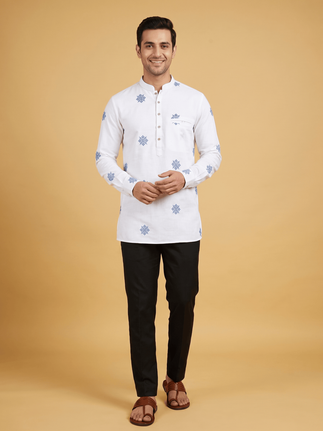 A man modeling a white long-sleeve kurta with light blue floral motifs, paired with black trousers and brown sandals, against a solid beige studio background. A man wearing an off-white cotton short kurta featuring blue geometric embroidery, styled with black trousers.