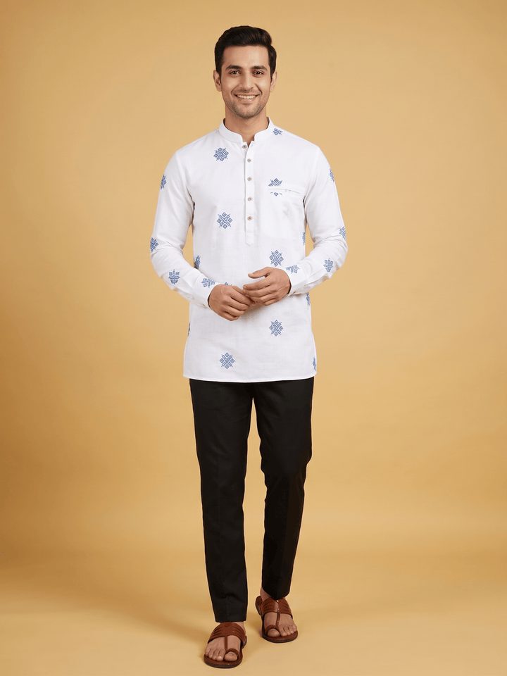 A man modeling a white long-sleeve kurta with light blue floral motifs, paired with black trousers and brown sandals, against a solid beige studio background. A man wearing an off-white cotton short kurta featuring blue geometric embroidery, styled with black trousers.