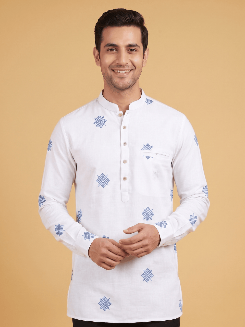 A man wearing a white long-sleeve kurta with blue geometric patterns, standing against a solid beige background. Close-up of the mandarin collar and blue geometric embroidery details on an off-white cotton short kurta.