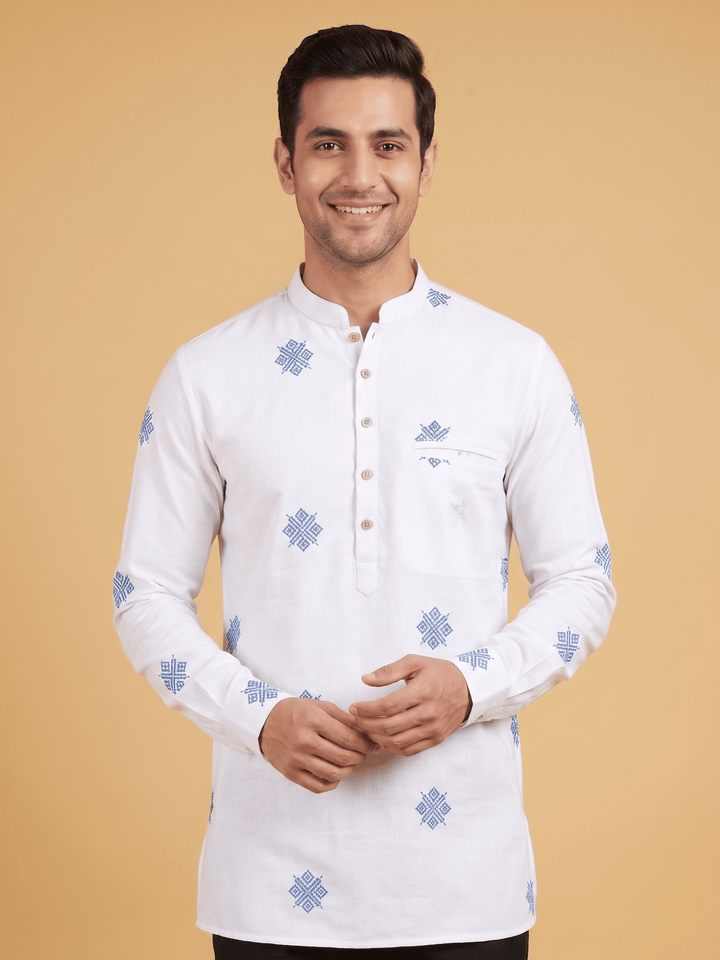A man wearing a white long-sleeve kurta with blue geometric patterns, standing against a solid beige background. Close-up of the mandarin collar and blue geometric embroidery details on an off-white cotton short kurta.