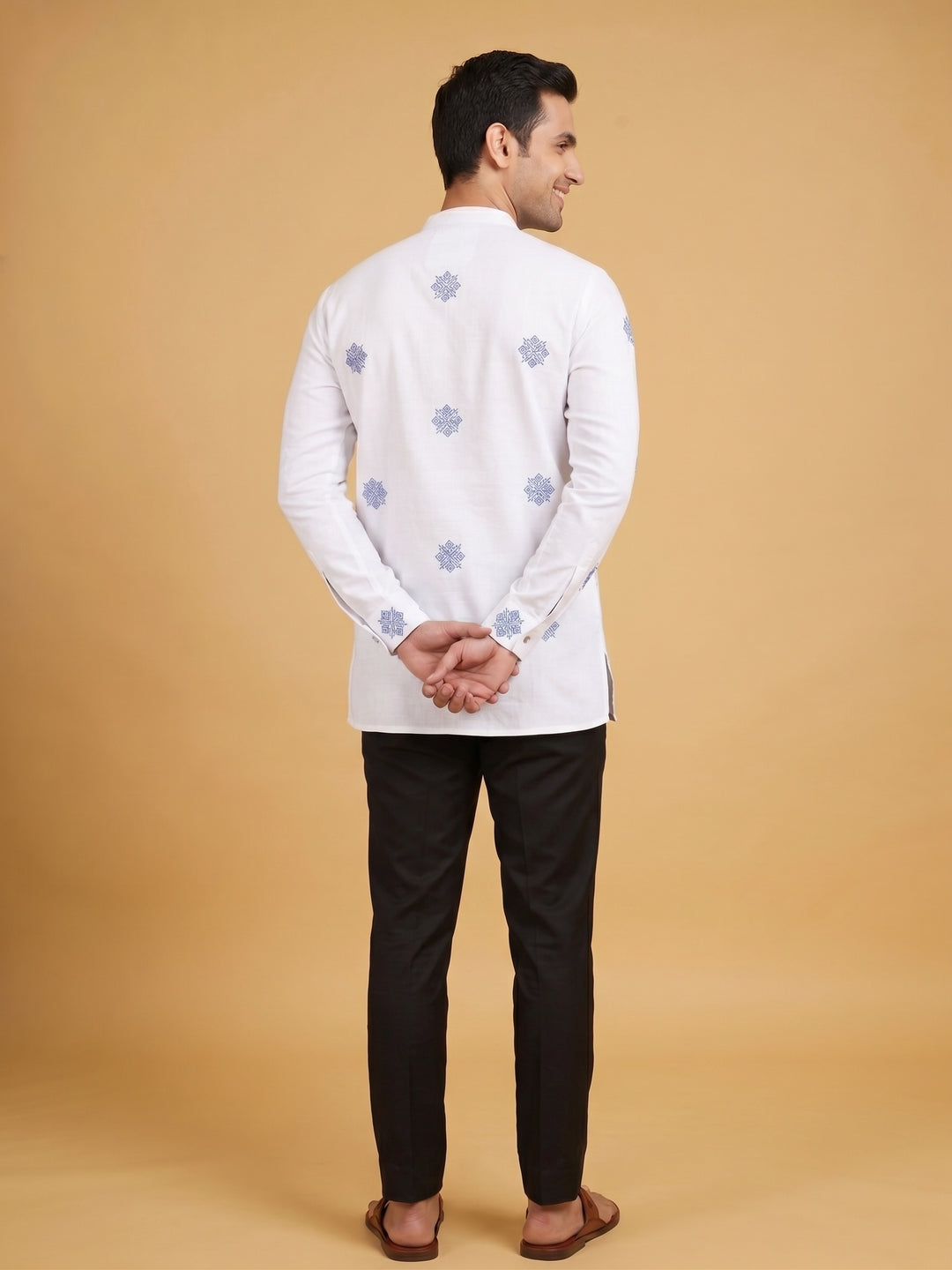 A man shown from the back wearing a white long-sleeve kurta with light blue floral embroidery, paired with black trousers and brown sandals, against a solid beige studio background. Back view of a man wearing an off-white cotton short kurta showcasing all-over blue geometric embroidery.