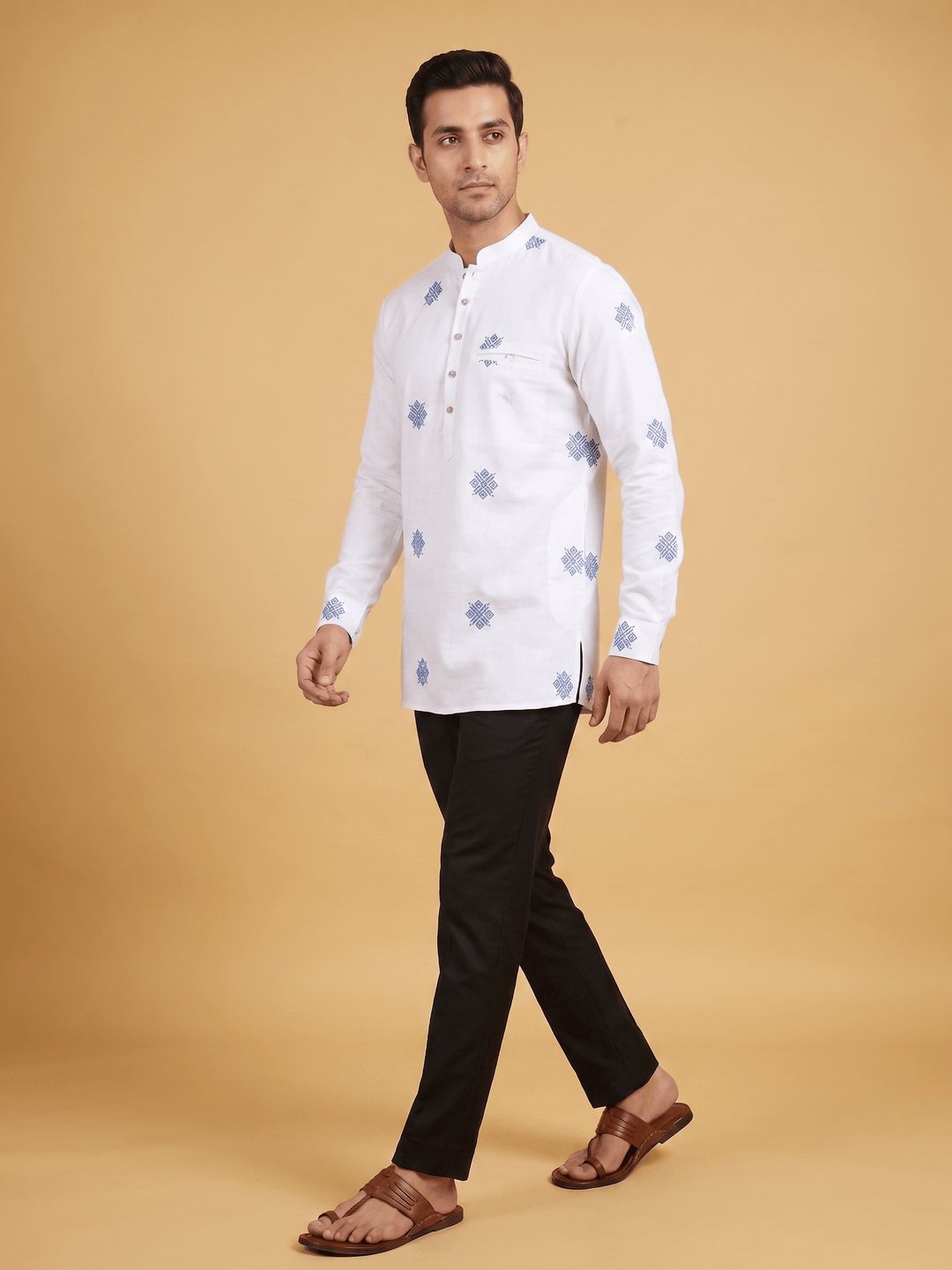 A man modeling a white long-sleeve kurta with light blue floral prints, paired with black trousers and brown sandals, against a solid beige studio background. Side-angle view of a man walking in an off-white cotton short kurta with blue geometric embroidery and black pants.