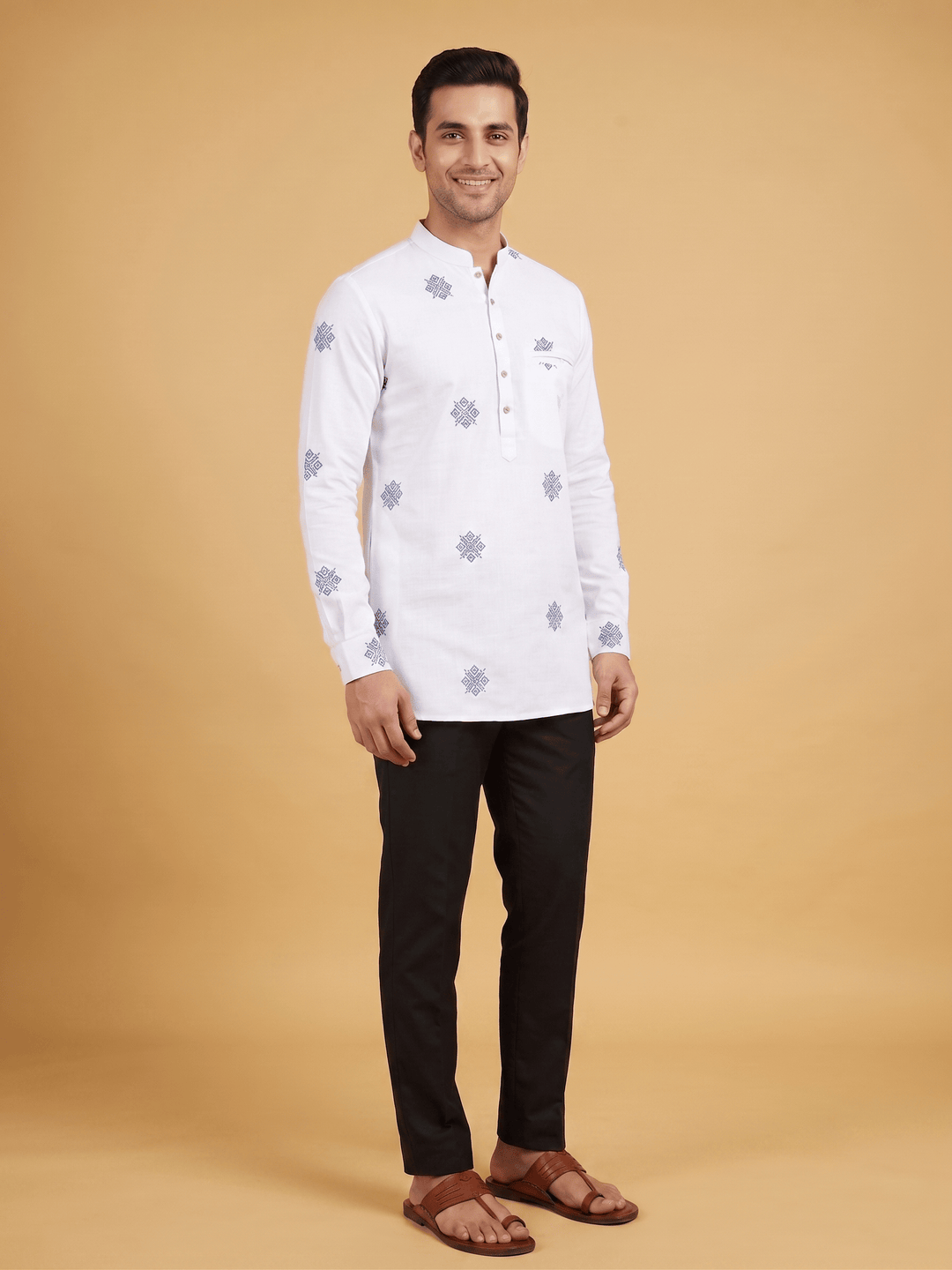 A man modeling a white long-sleeve kurta with light blue floral patterns, paired with black trousers and brown sandals, against a solid beige studio background. Full-length front view of a man modeling an off-white cotton short kurta with blue geometric patterns and black trousers.