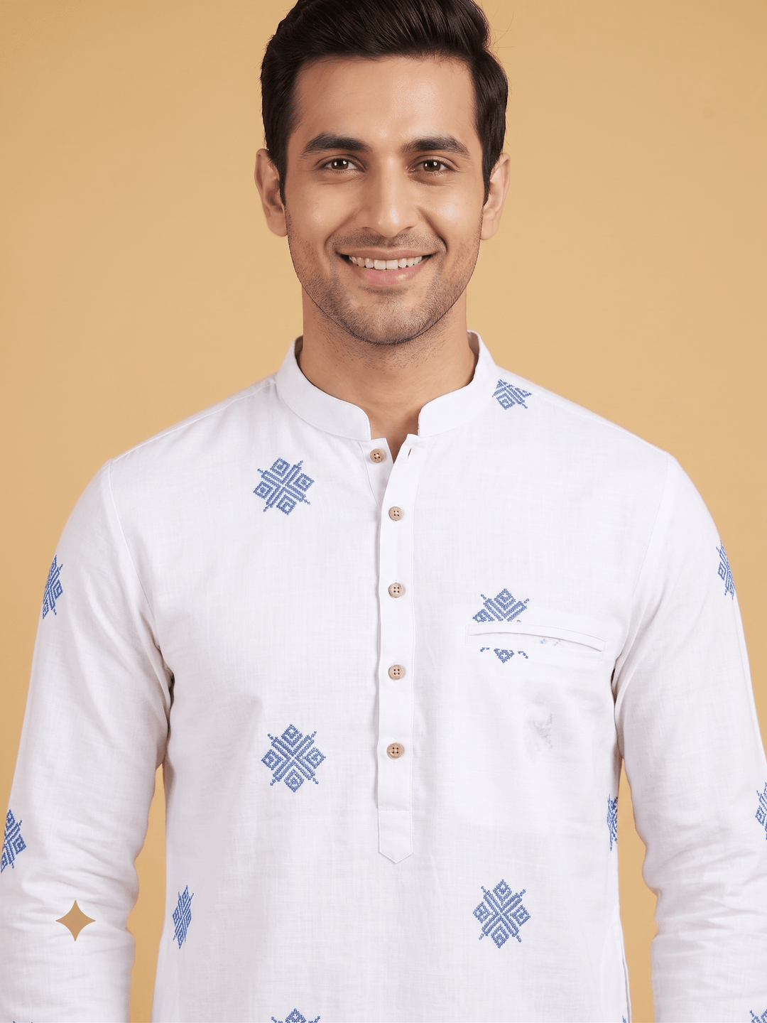 A smiling man wearing a white long-sleeve kurta with blue geometric patterns and a buttoned mandarin collar, set against a solid beige background. Medium close-up shot of a man wearing an off-white cotton short kurta with blue geometric embroidery and a buttoned placket.