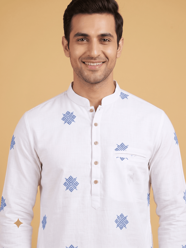 A smiling man wearing a white long-sleeve kurta with blue geometric patterns and a buttoned mandarin collar, set against a solid beige background. Medium close-up shot of a man wearing an off-white cotton short kurta with blue geometric embroidery and a buttoned placket.