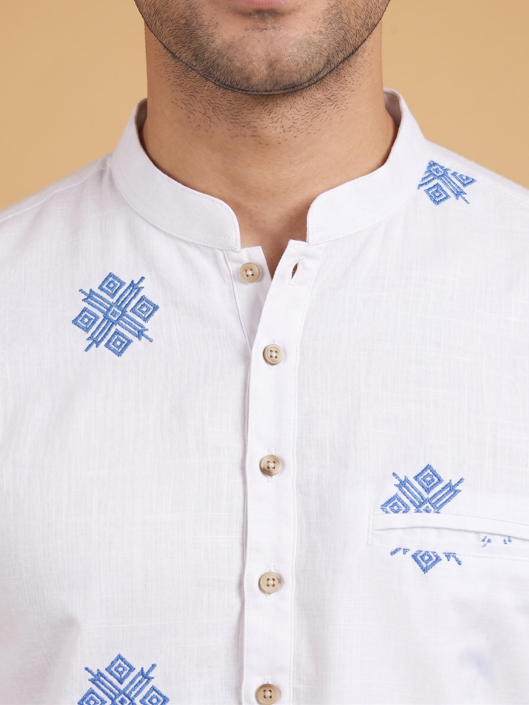 Close-up of a man wearing a white mandarin-collar button-up shirt with blue geometric embroidery on the chest and collar, set against a solid beige background. Front-facing medium shot of a man wearing an off-white cotton short kurta with blue geometric embroidery. Close-up of the mandarin collar and blue geometric embroidery details on an off-white cotton short kurta.