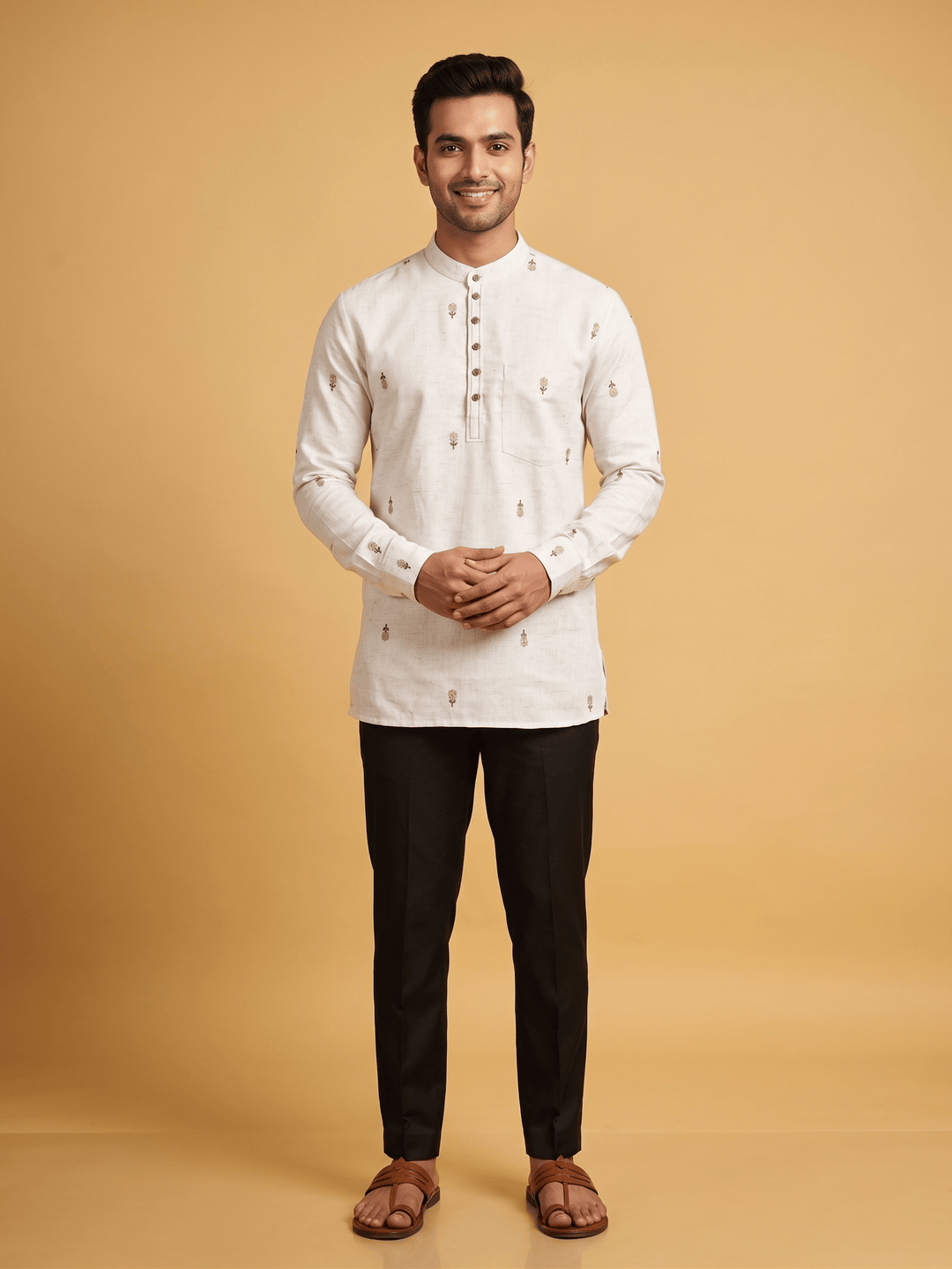 A man modeling a white long-sleeve kurta with small brown embroidered motifs, paired with black trousers and brown sandals, against a solid warm beige background. A man walking in an off-white cotton short kurta with minimalist floral embroidery, paired with black trousers.