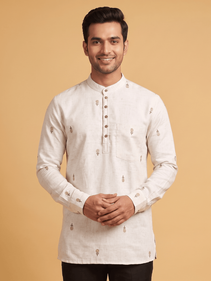 A man wearing a light beige long-sleeve kurta with small embroidered motifs, standing against a solid warm beige background. Close-up of the mandarin collar and subtle floral embroidery on an off-white cotton short kurta.