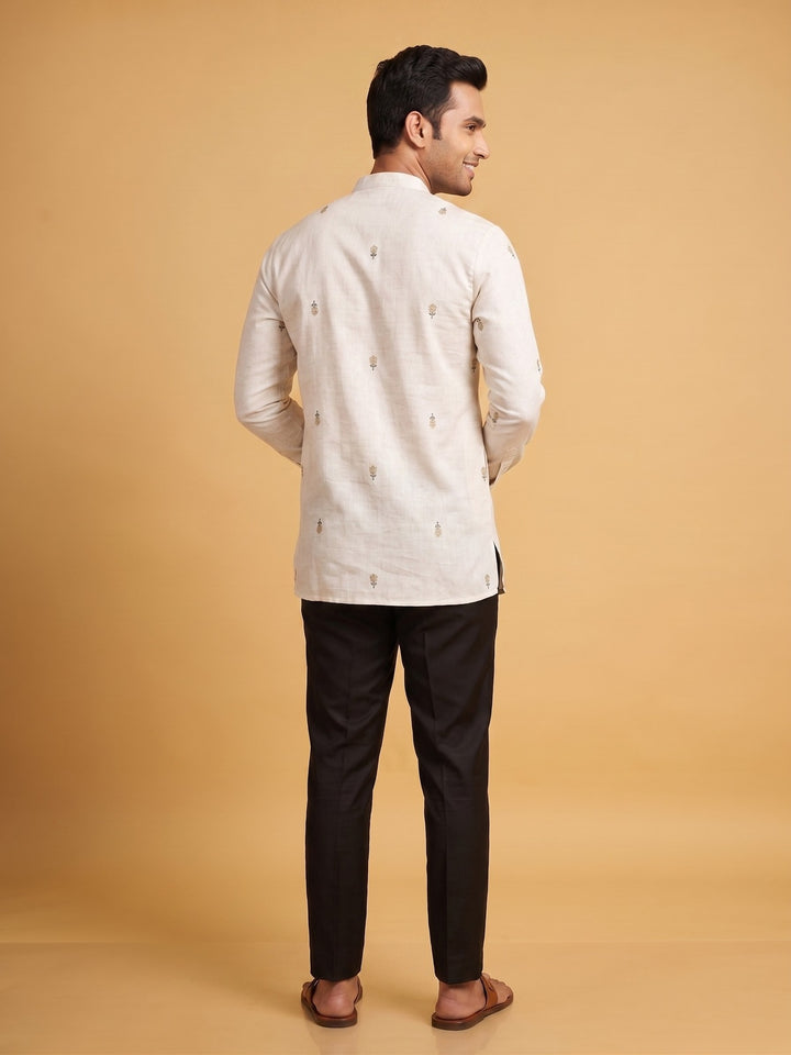 A man shown from the back wearing a light beige kurta with small embroidered details and black trousers, standing against a solid warm beige studio background. Back view of a man wearing an off-white cotton short kurta featuring all-over minimalist floral embroidery.